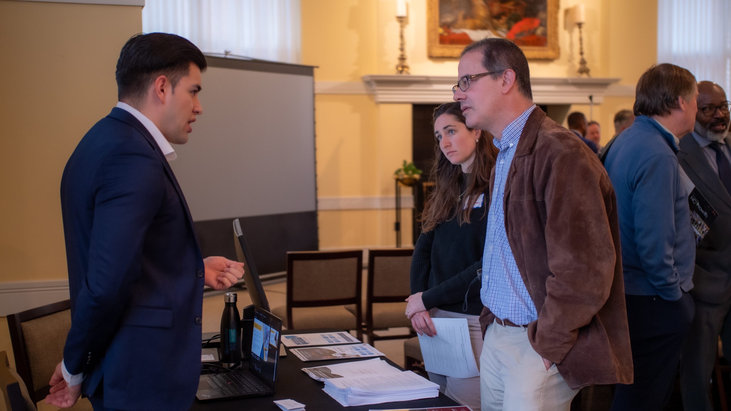 People converse at a table with papers and a laptop during an indoor event.