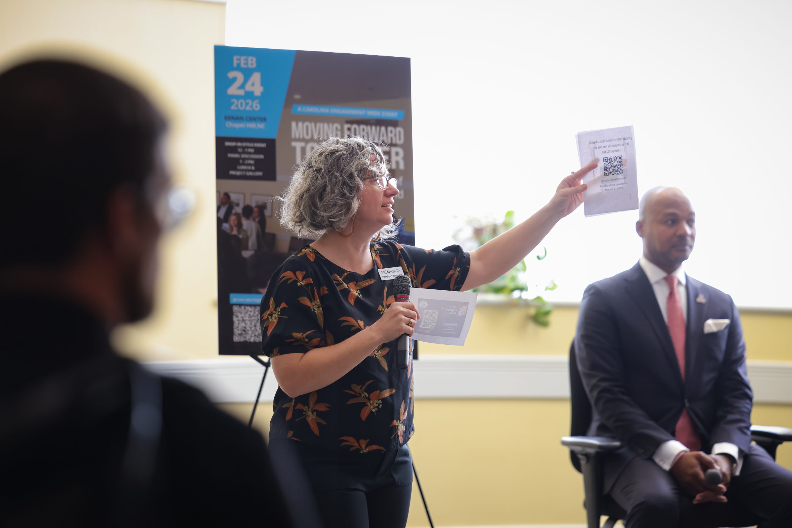 A woman with curly gray hair wearing glasses and a floral print shirt holds a flyer with a QR code and a microphone, speaking to an audience. A man in a suit and tie is seated to her right. A poster with "FEB 24 2026" and "MOVING FORWARD TOGETHER" is in the background.