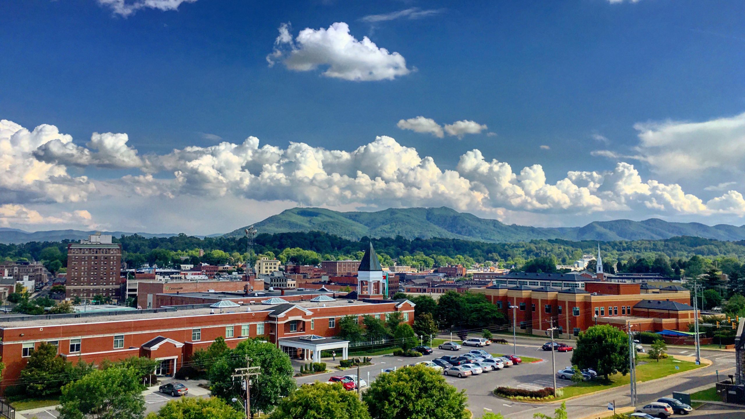 Aerial view of a brick building complex with a parking lot in the foreground, and a town with mountains in the background under a blue sky with white clouds.