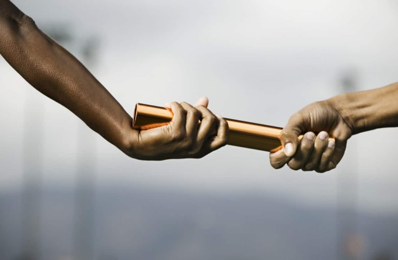 Close-up of two hands, one with darker skin and one with lighter skin, passing a bronze baton during a race.