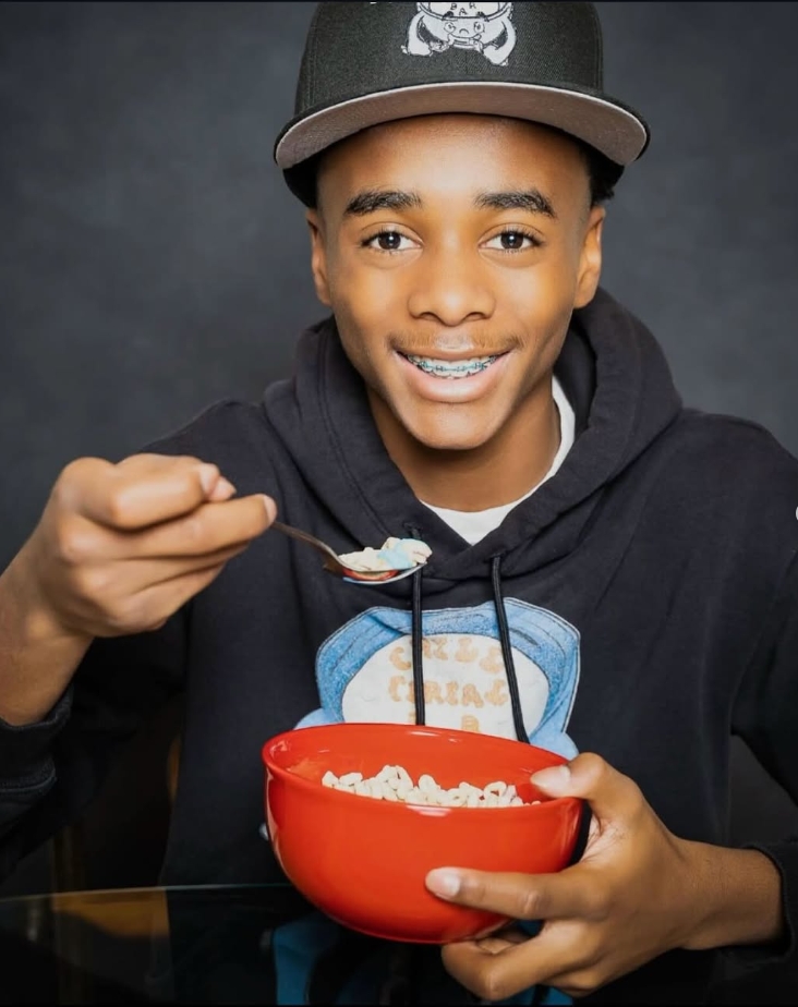 A young person wearing a black baseball cap and a black hoodie smiles while eating cereal from a red bowl with a spoon.