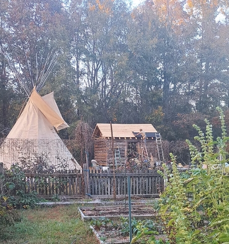 A teepee stands next to a wooden structure being built, with people working on its roof.