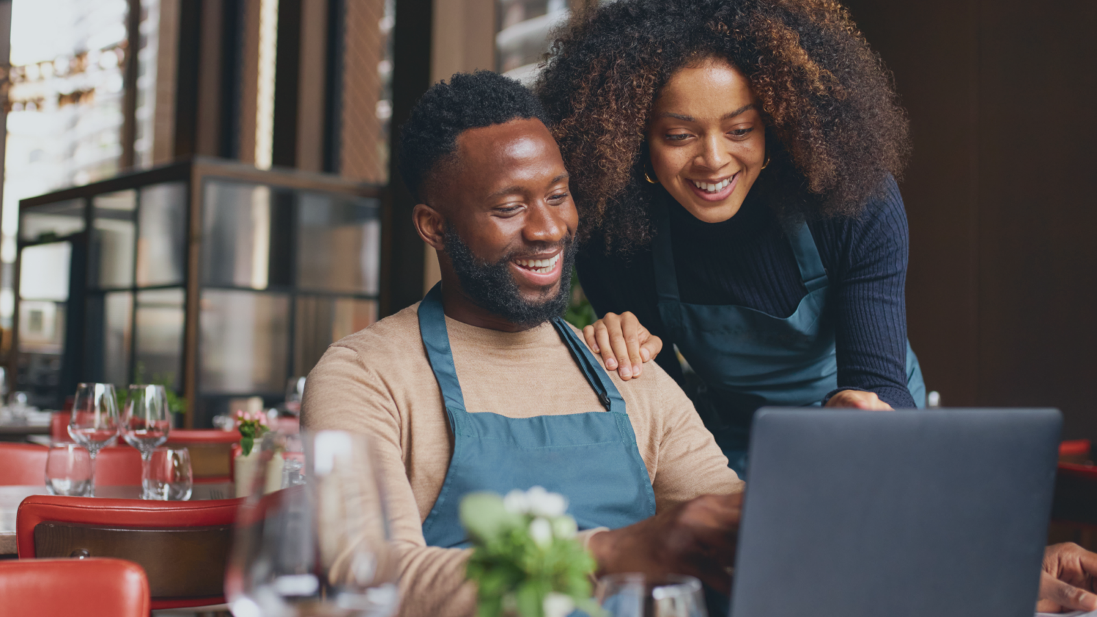 Two restaurant staff members wearing aprons look at a laptop together at a table in a restaurant.