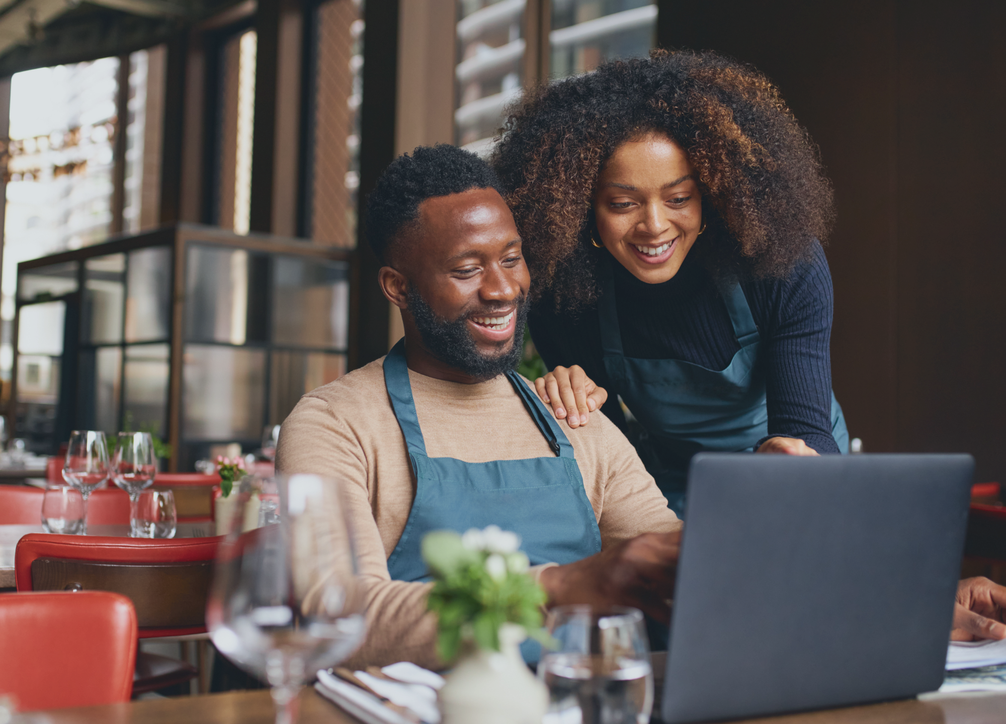 Two restaurant staff members wearing aprons look at a laptop together at a table in a restaurant.