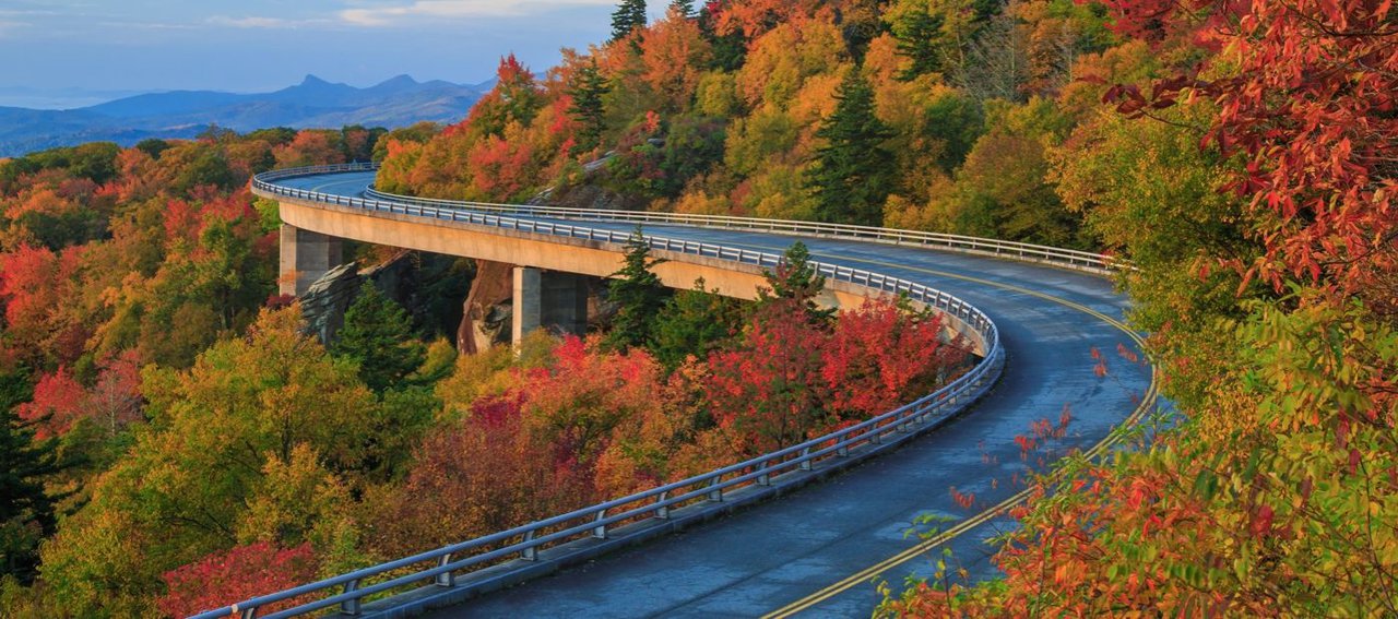 A winding road curves over an elevated bridge, surrounded by a vibrant autumn forest with trees in shades of red, orange, and yellow.